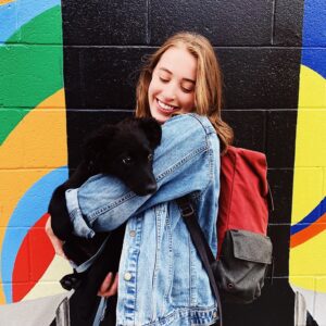 A white woman with blonde hair, Ciara, holds a small black puppy, Eddie, in front of a colorful background.