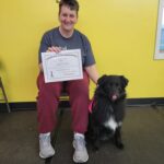 Denise, wearing red pants and t-shirt, sits in a chair and smiles while holding up a graduation certificate for her black Australian Shepherd, Lexi. 
