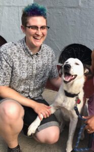 Jaime, a smiling person with blue hair and glasses, kneels down next to their dog Astro. 