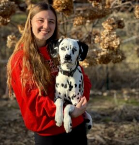 Chesney, wearing a red sweatshirt with long blond hair, smiles at the camera while holding her Dalmatian puppy, Bishop.