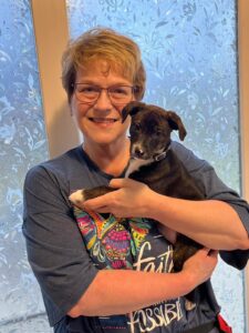 Michelle, a woman with light hair in a short cut, glasses, and a big smile, poses with her brindle and white puppy, Whimsy.